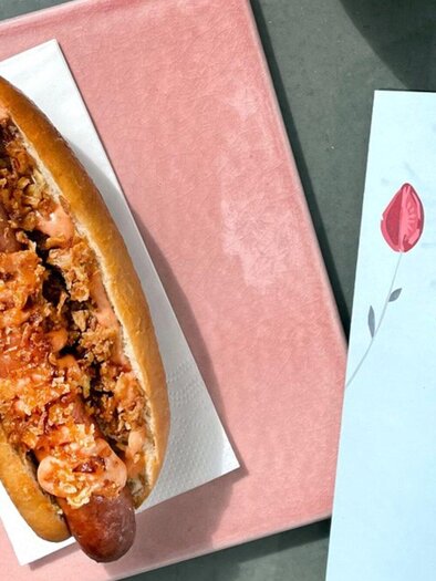 A hotdog with tender sausage and crispy toppings lies on a pink plate. Next to it is a menu with colorful floral motifs. | © Salaettl