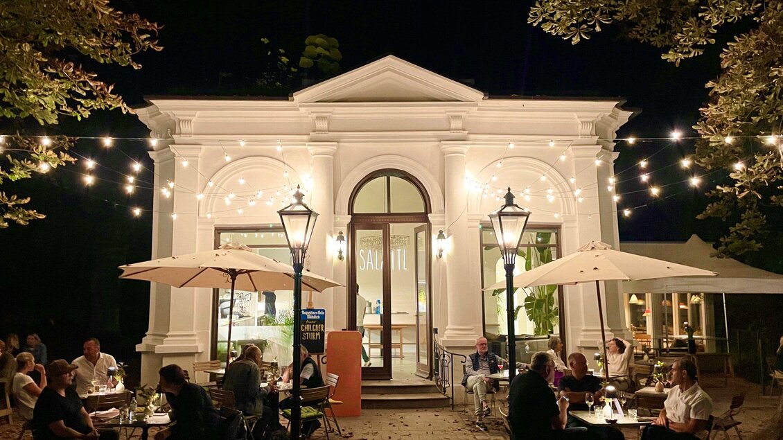 Eine gemütliche Restaurantterrasse bei Nacht mit beleuchteten Laternen und mehreren Tischen. Das historische Gebäude ist einladend und von Bäumen umgeben. | © Salaettl