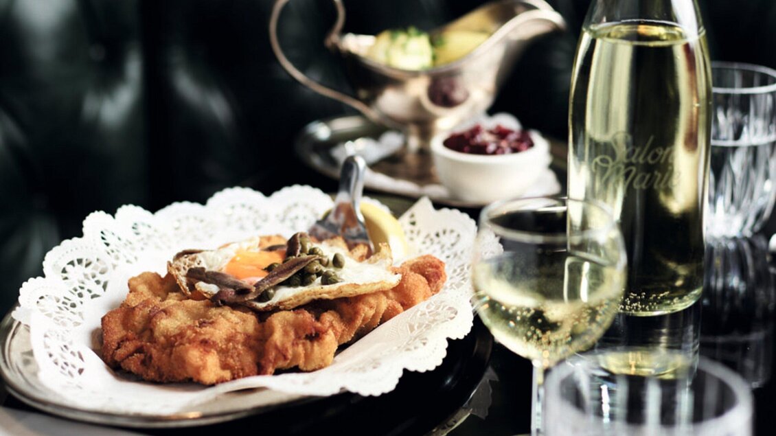 Ein saftiges Schnitzel, serviert mit einem Spiegelei und verschiedenen Beilagen. Im Hintergrund stehen ein Glas Weißwein und eine Flasche Wasser. | © Salon Marie