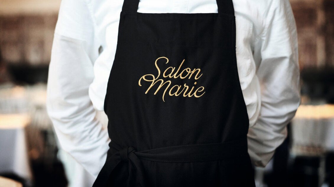 Ein Kellner trägt eine schwarze Schürze mit der goldenen Aufschrift "Salon Marie". Im Hintergrund sind Tische in einem eleganten Restaurant zu sehen. | © Salon Marie
