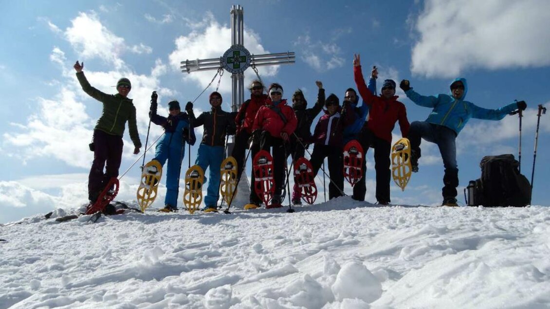 Eine Gruppe von Menschen steht glücklich auf einem schneebedeckten Gipfel. Im Hintergrund ist ein Kreuz und ein klarer Himmel zu sehen. | © Salzstieglhaus