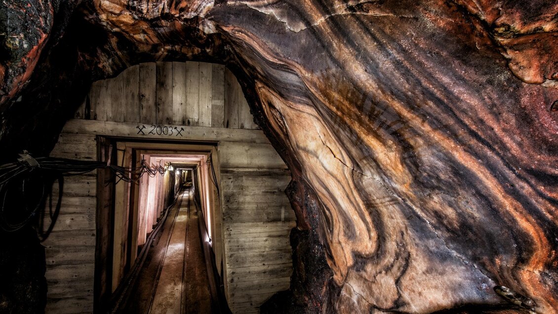 Ein schmaler Tunnel mit holzverkleideten Wänden und buntem Gestein. Der Weg verläuft in die Dunkelheit. | © Salzwelten/Kernmayer