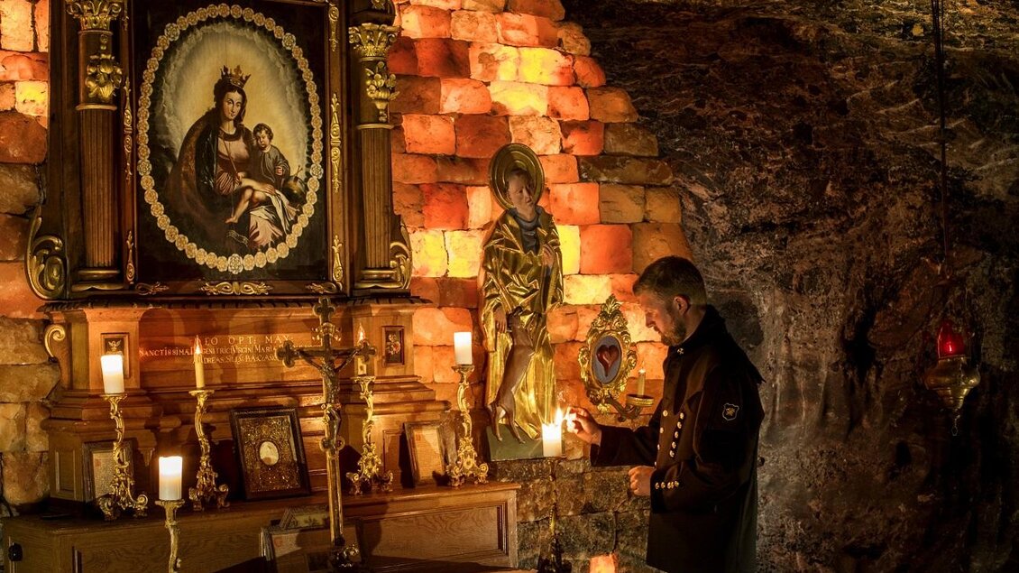 Ein Mann steht in einer Höhle vor einem Altar mit religiösen Bildern. Sanftes Licht beleuchtet die Wände und Kerzen. | © Salzwelten/Kernmayer