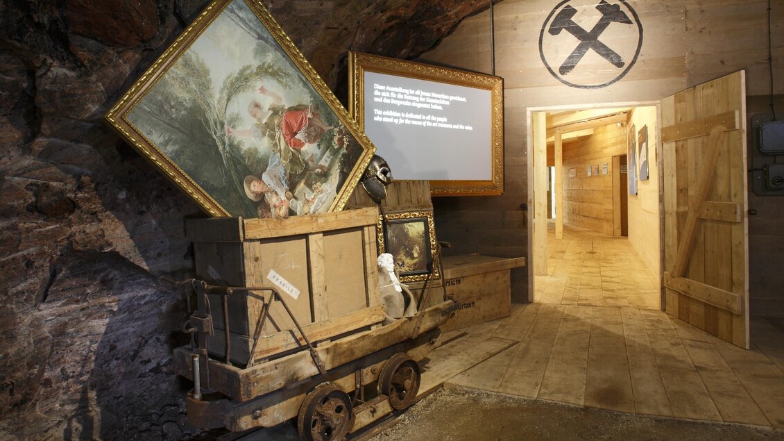 Ein Kunstwerk in einem alten Bergwerk mit einer Holzkarren und einer Eingangstür. Im Hintergrund sieht man ein Symbol für den Bergbau. | © Salzwelten/Mario Schmid