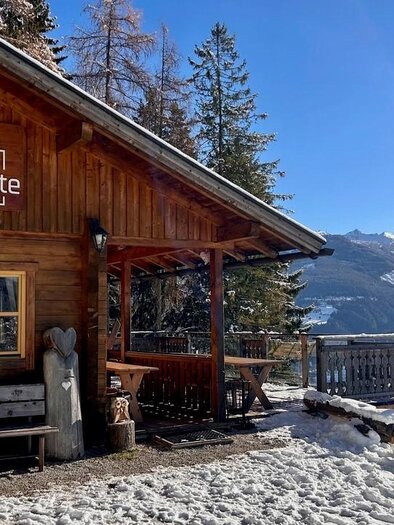 Eine gemütliche Hütte aus Holz mit dem Schild "Sattelberghütte". Im Hintergrund sind schneebedeckte Berge und ein blauer Himmel zu sehen. | © Sattelberghgütte