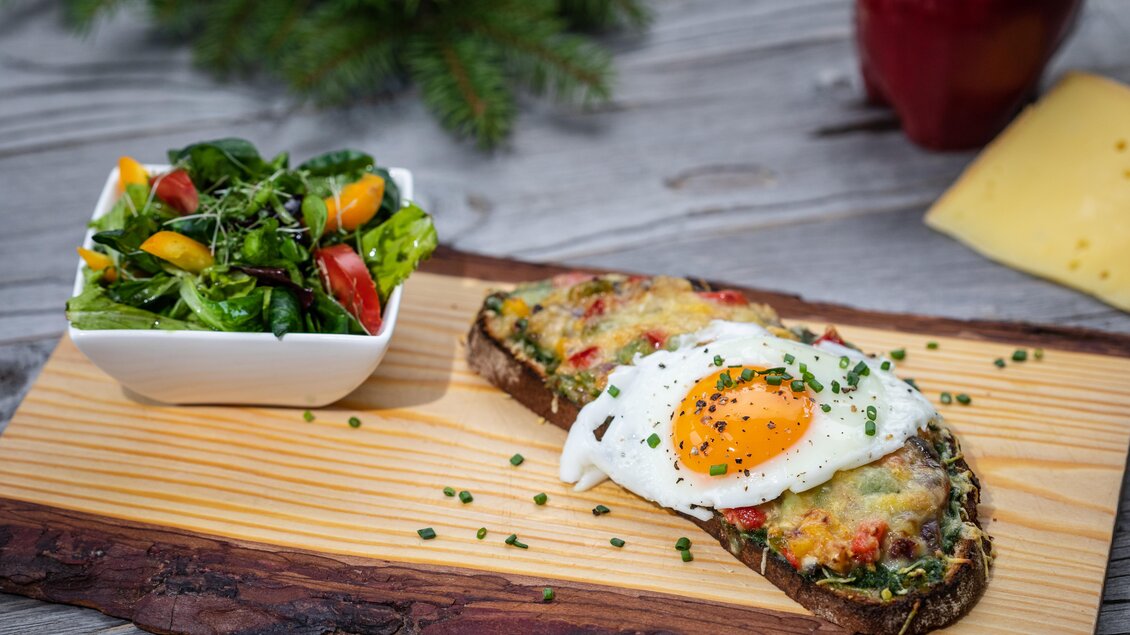 Ein köstliches Gericht mit einem belegten Brot, einem Spiegelei und Käse, serviert auf einem Holzbrett. Dazu gibt es einen frischen Salat in einer kleinen Schüssel. | © Martin Huber