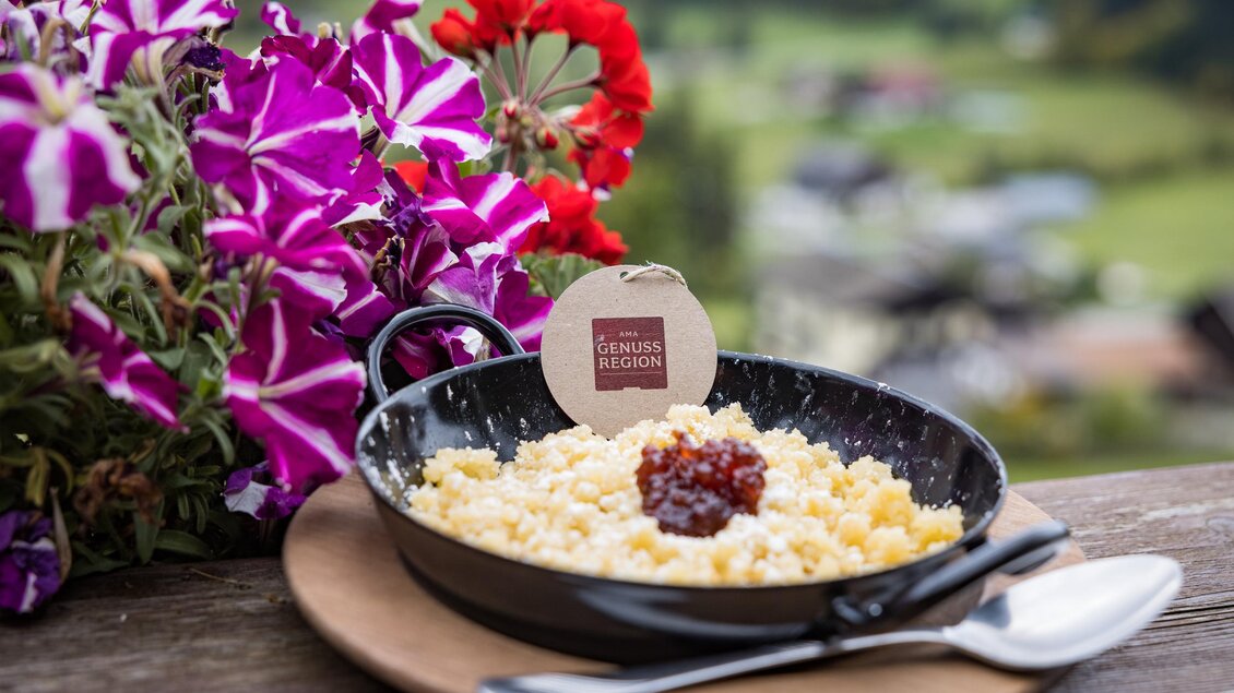 Ein Gericht mit Knödeln, serviert in einer schwarzen Schale, dekoriert mit einer roten Fruchtsoße. Im Hintergrund sind bunte Blumen und eine grüne Landschaft sichtbar. | © Netzwerk Kulinarik wildbild