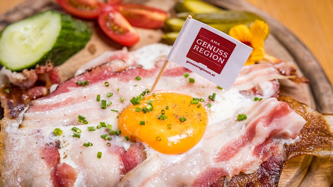 Ein herzhaftes Gericht mit Speck, einem frischen Eigelb und garnierenden Zutaten wie Tomaten und Gurken. Auf dem Teller befindet sich eine kleine Fahne mit dem Aufdruck „Genussregion“. | © Netzwerk Kulinarik wildbild
