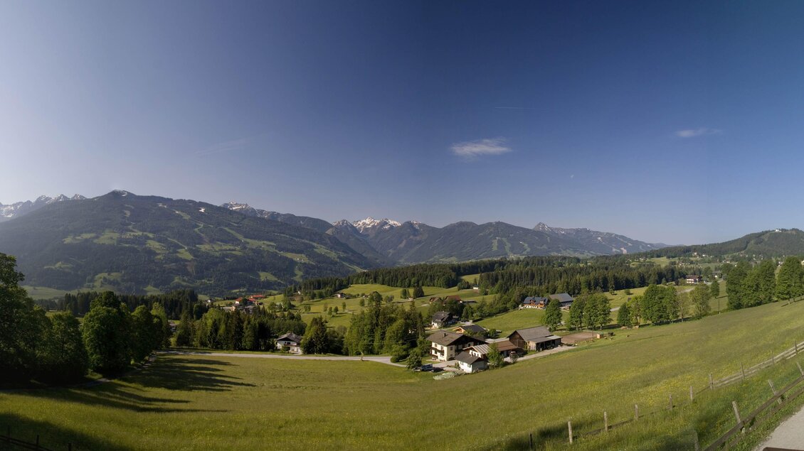 Eine malerische Landschaft mit grünen Wiesen und kleinen Häusern. Im Hintergrund ragen majestätische Berge und ein klarer Himmel hervor. | © Sattelberghütte