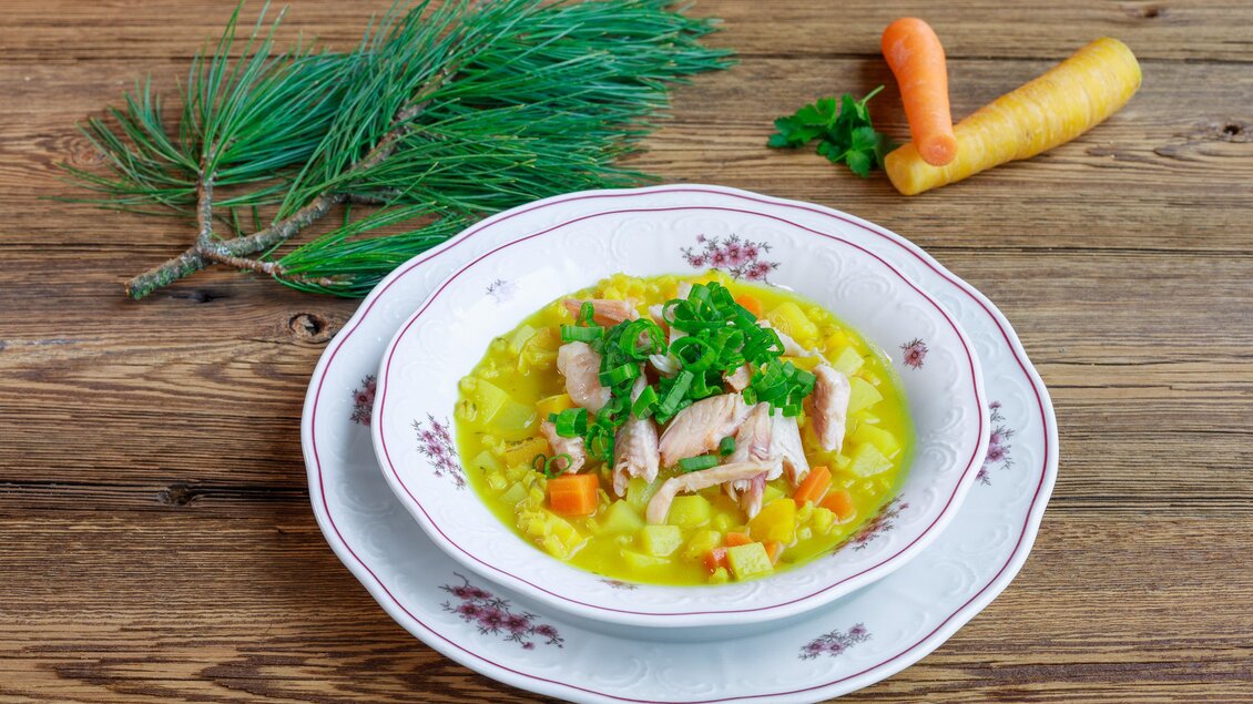 Eine köstliche Suppe mit Hühnerfleisch, Gemüse und Kräutern in einer weißen Schale. Im Hintergrund sind frische Karotten und Tannenzweige zu sehen. | © Martin Huber