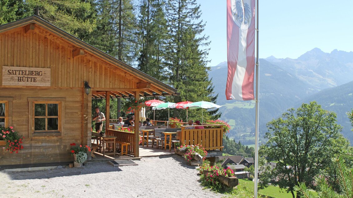 Eine gemütliche Berghütte mit einer Terrasse, umgeben von Bäumen und Bergen. Auf der Terrasse genießen Gäste das schöne Wetter. | © Sattelberghütte