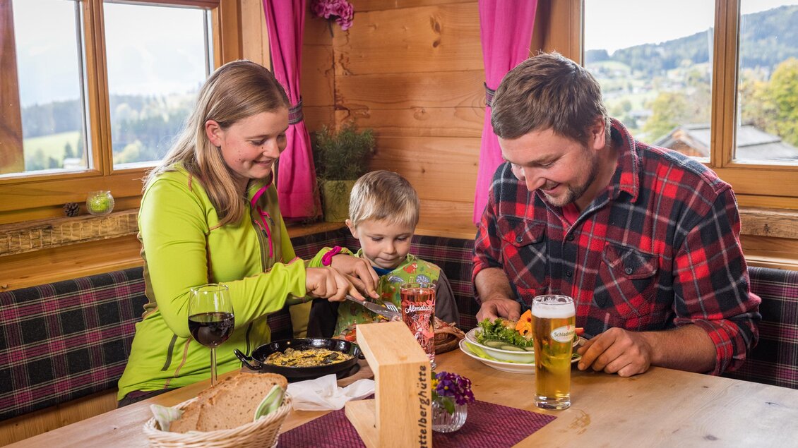 Eine Familie sitzt an einem Tisch in einer Hütte und genießt eine Mahlzeit zusammen. Es gibt Essen, Getränke und eine gemütliche Atmosphäre mit Blick auf die Landschaft. | © Netzwerk Kulinarik wildbild