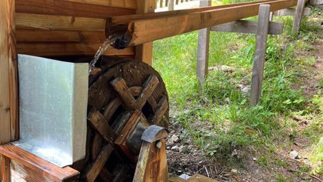 Ein altes Wasserrad aus Holz, das an einem kleinen Gebäude befestigt ist. Der Bereich ist von Grünflächen und Bäumen umgeben. | © Sattelberghütte