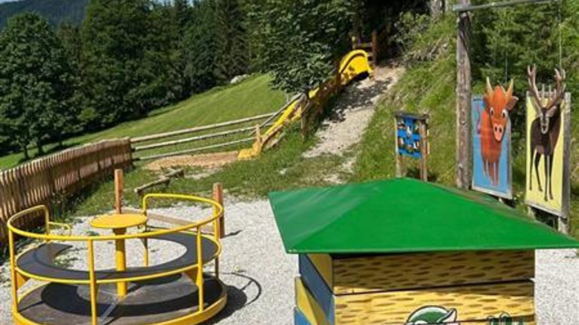 Ein Spielplatz in der Natur mit einem Karrussell und einer Rutsche. Im Hintergrund sind Bäume und blauer Himmel zu sehen. | © Sattelberghütte