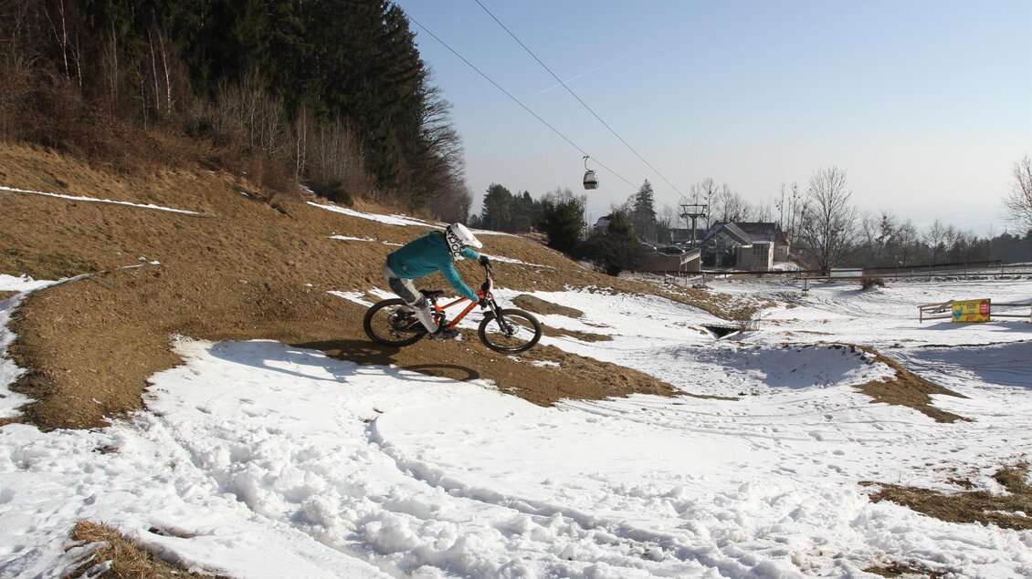 Ein Mountainbiker fährt auf einem schneebedeckten Gelände. Im Hintergrund sind Bäume und eine Seilbahn sichtbar. | © The Gap Coaching