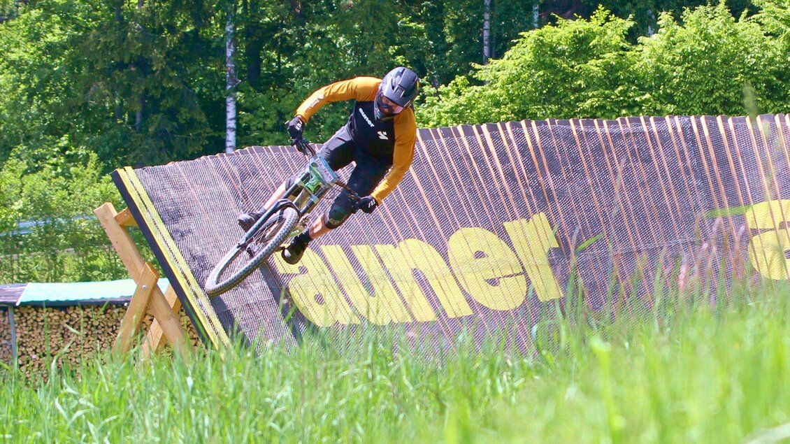 Ein Mountainbiker fährt eine Rampe und macht einen Sprung. Im Hintergrund sind Bäume und eine grüne Wiese zu sehen.