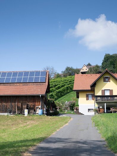 A rural landscape with two buildings, one with solar panels on the roof. In the background, there are gentle hills and a blue sky. | © TV Südsteiermark - Dominik Feibel