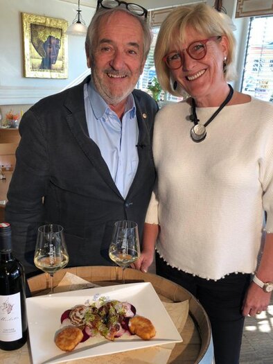 A man and a woman are smiling in front of a table decoration with a plate of food and two wine glasses. In the background, a cozy restaurant atmosphere can be seen. | © Schaller's Gastwirtschaft