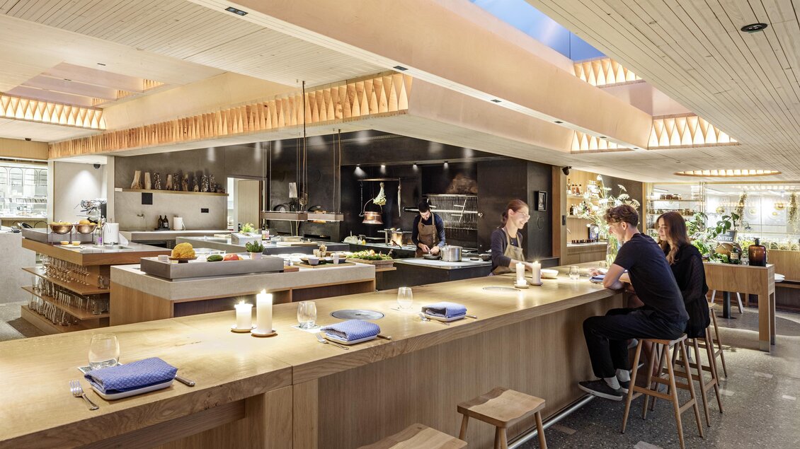 Ein modernes Restaurant mit einer offenen Küche und einer Holztheke. Gäste sitzen im Hintergrund und genießen ihre Mahlzeiten in einem einladenden Ambiente. | © Restaurant Steirereck/Hertha Hurnaus
