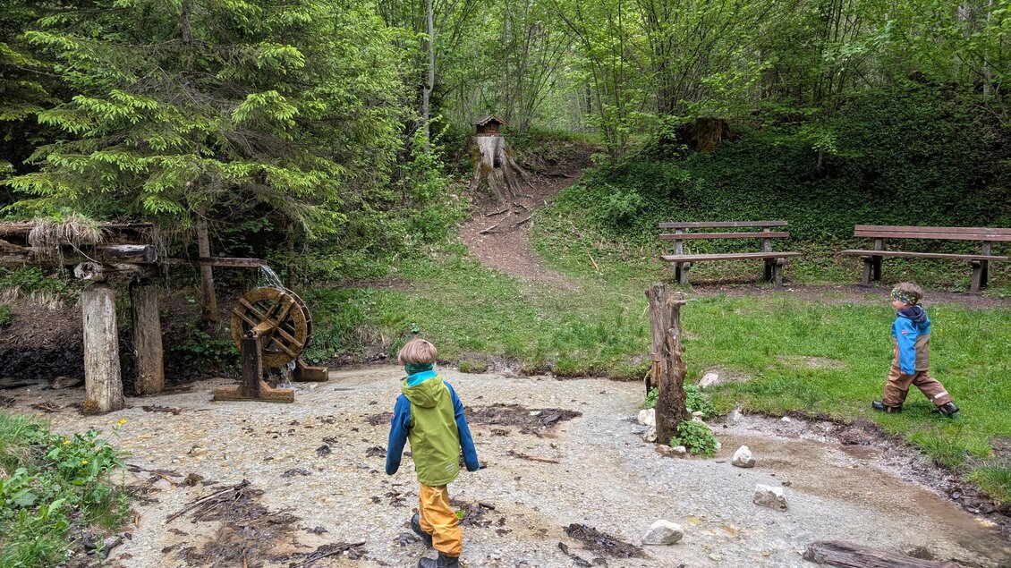 Zwei Kinder spielen in einem Waldgebiet mit einer Wassermühle. Die Umgebung ist grün und einladend mit Bänken zum Sitzen. | © Stefan Wieser-Bliem