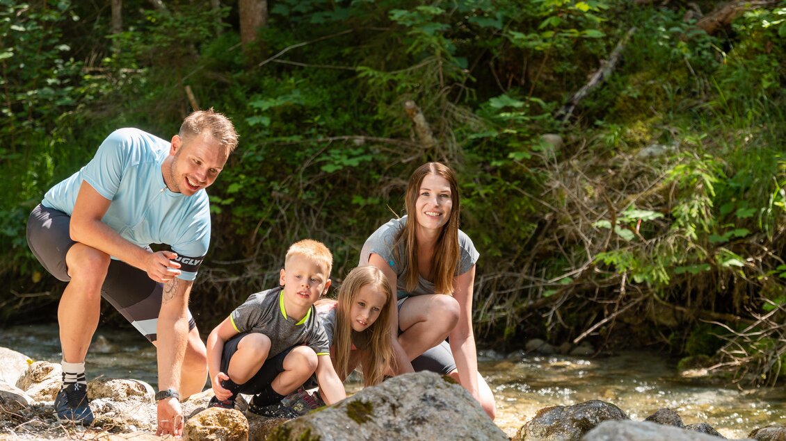 Eine Familie erkundet einen klaren Bach in der Natur. Die Kinder spielen im Wasser, während die Erwachsenen beobachten und Freude haben. | © Lorenz Maser