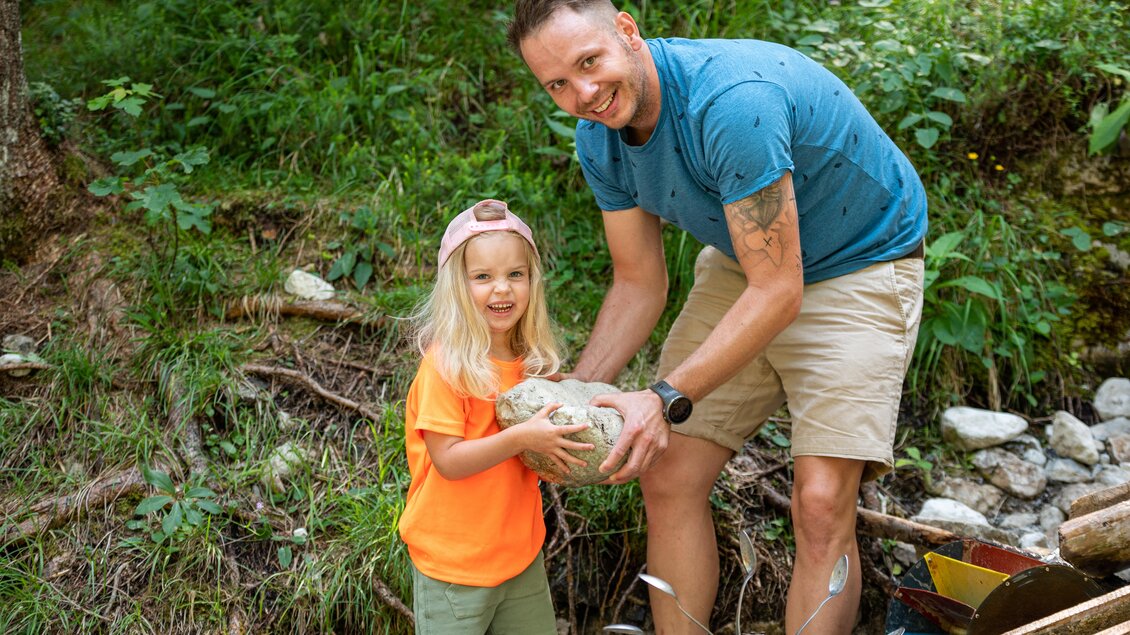 Ein Vater und seine Tochter heben gemeinsam einen großen Stein an. Im Hintergrund sind grüne Bäume und eine natürliche Umgebung zu sehen. | © Lorenz Maser 