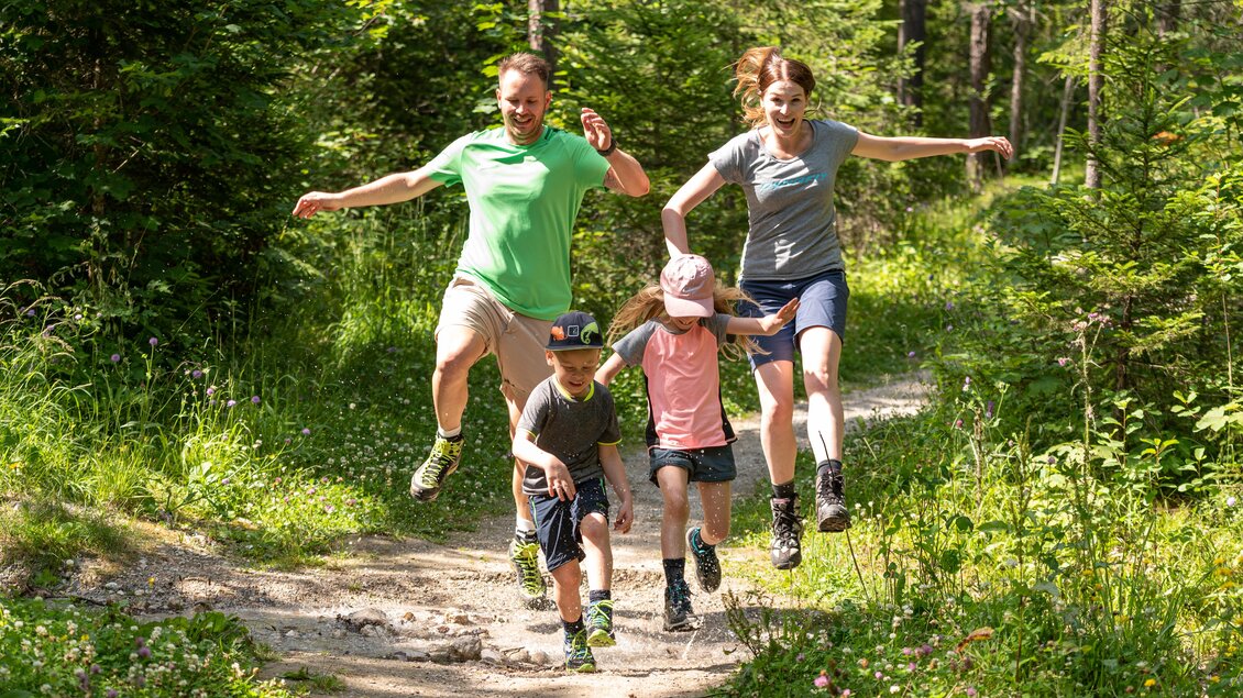 Eine Familie mit zwei Erwachsenen und zwei Kindern springt fröhlich auf einem Waldweg. Die Sonne scheint und die Umgebung ist grün und lebendig. | © Lorenz Maser 