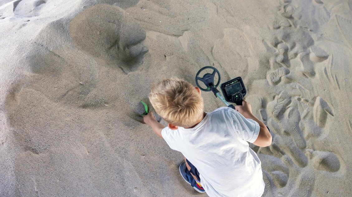 Ein Junge sucht im Sand mit einem Metalldetektor. Er wirkt konzentriert und spielt im Freien. | © www.zloam.at