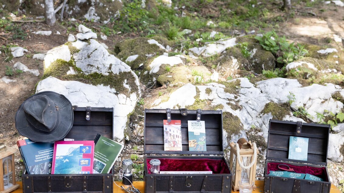 Drei offene Schatztruhen stehen auf einem Tisch im Freien. In ihnen befinden sich Bücher, ein Hut und verschiedene Schriftstücke, umgeben von Natur. | © www.zloam.at