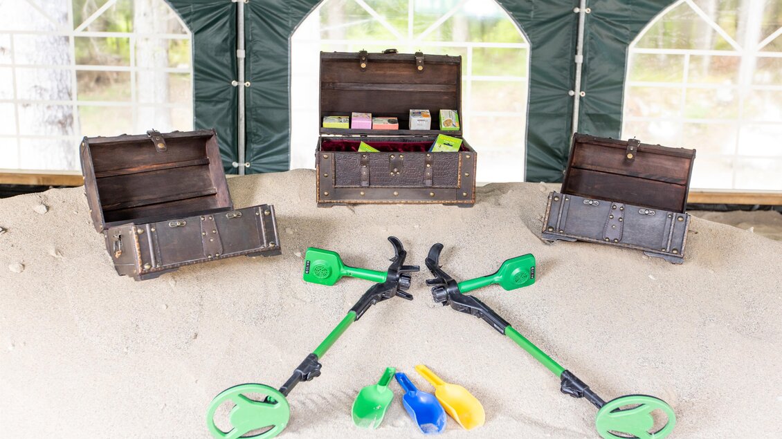 Zwei Kisten stehen auf einem Sandhaufen, gefüllt mit verschiedenen Gegenständen. Darum liegen zwei Metallldetektoren und Schaufeln in bunten Farben. | © www.zloam.at