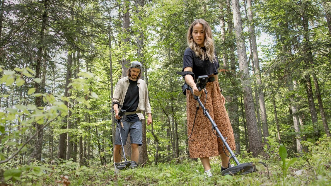 Zwei Personen suchen mit Metalldetektoren in einem Waldgebiet. Die Umgebung ist grün und sonnig, umgeben von hohen Bäumen. | © www.zloam.at