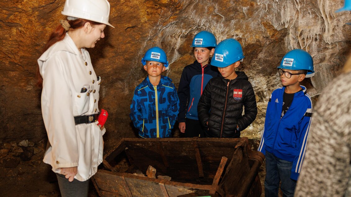Eine Gruppen von Kindern mit blauen Helmen steht in einer Höhle und hört einer Anleitung zu. Eine Person in weißer Kleidung zeigt auf ein altes Holzgestell. | © Anita Fössl