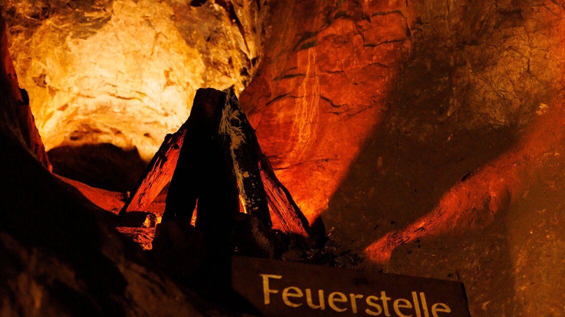 Eine gemütliche Feuerstelle in einer Höhle mit warmem Licht und rustikalem Umfeld. Der Namenstafel „Feuerstelle“ ist deutlich sichtbar. | © Anita Fössl