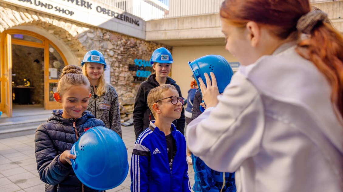 Eine Gruppe von Kindern steht vor einem Museum und schaut auf einen Helm. Einige Kinder tragen bereits blaue Helme und scheinen aufgeregt zu sein. | © Anita Fössl