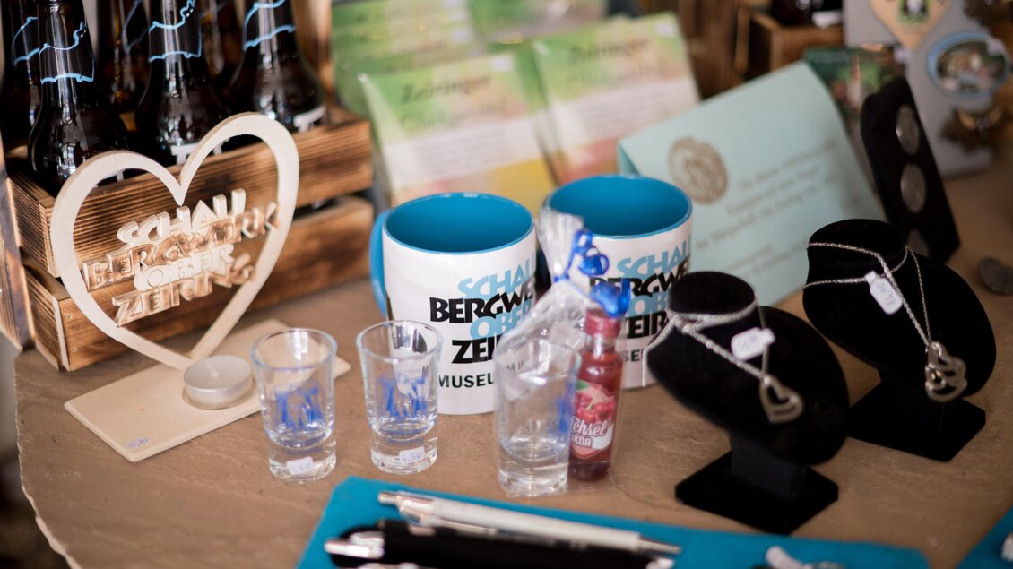 Ein Tisch mit Souvenirs, darunter Tassen, Gläser und Schmuck. Zudem sind einige Flaschen und Stifte sichtbar, die zum Angebot gehören. | © Anita Fössl