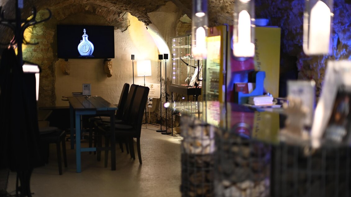 Ein gemütlicher Raum mit einer modernen Einrichtung und einer stimmungsvollen Beleuchtung. Im Hintergrund sind Ausstellungsgegenstände und ein Bildschirm sichtbar. | © Anita Fössl