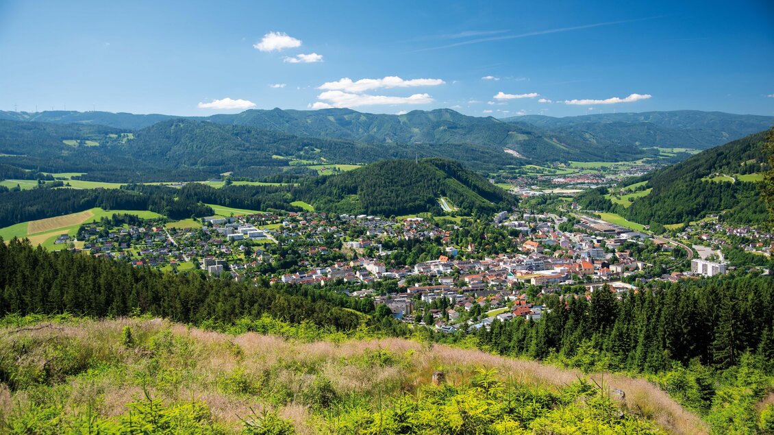 Eine malerische Aussicht auf eine kleine Stadt umgeben von grünen Hügeln und Bergen. Der Himmel ist klar und blau mit einigen Wolken.