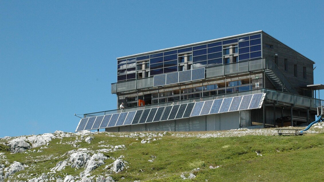 Ein modernes Gebäude auf einem Hügel mit großen Fenstern und Solarpanelen. Der Himmel ist klar und blau, umgeben von einer grünen Wiese.