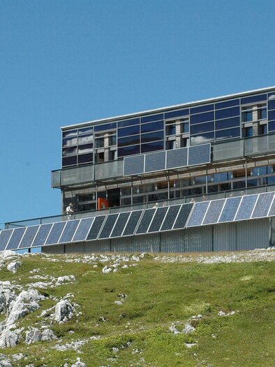 Ein modernes Gebäude auf einem Hügel mit großen Fenstern und Solarpanelen. Der Himmel ist klar und blau, umgeben von einer grünen Wiese.