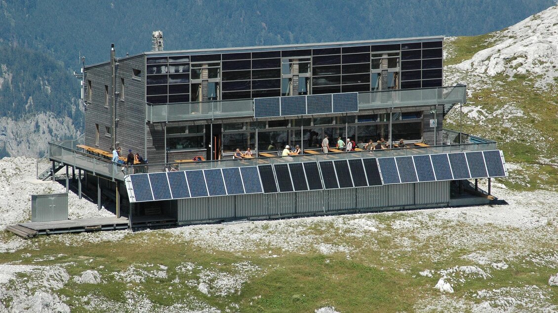 Ein modernes Gebäude auf einer Bergspitze, umgeben von felsigem Gelände. Das Gebäude hat große Fenster und ist mit Solarpanelen ausgestattet.