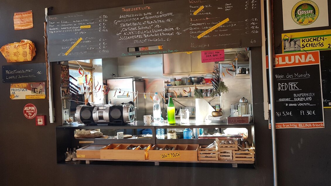 Ein kleines, gemütliches Café mit einer offen gestalteten Theke und einer Auswahl an Getränken. An der Wand hängen Schrifttafeln mit dem Menü und Angeboten.