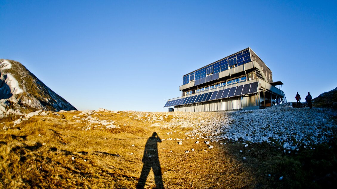 Ein modernes Gebäude in einer Berglandschaft mit klarem Himmel. Im Hintergrund sind Hügel und Felsen zu sehen.