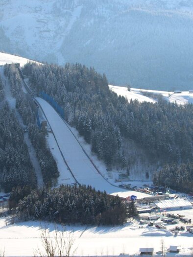 Eine schneebedeckte Landschaft mit einem Sprungschanzenkomplex vor einem bewaldeten Hügel. Im Hintergrund sind Berge und ein klarer Himmel zu sehen. | © TVB Ausseerland Salzkammergut/Kolb