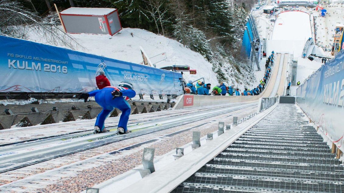 Eine Skisprungschanze im verschneiten Kulm. Sportler bereiten sich auf den Sprung vor, während Zuschauer und Veranstaltungsbanner im Hintergrund zu sehen sind. | © Austria Ski Nordic Veranstaltungs GmbH