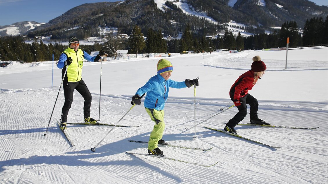 Eine Familie genießt das Langlaufen im Schnee. Im Hintergrund sind schneebedeckte Berge und eine klare, sonnige Winterlandschaft zu sehen. | © Erlebnisregion Murtal