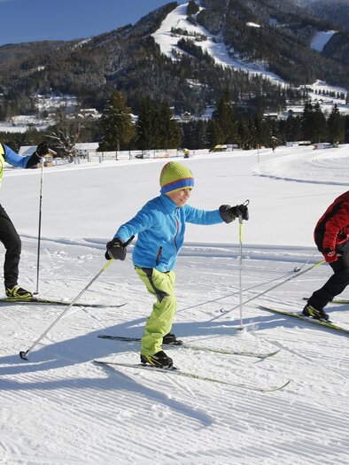 A family enjoys cross-country skiing in the snow. In the background, snow-covered mountains and a clear, sunny winter landscape can be seen. | © Erlebnisregion Murtal