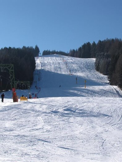 A snow-covered slope under a clear blue sky. In the background, tall trees rise up and a ski lift is visible. | © Skilift Obdach