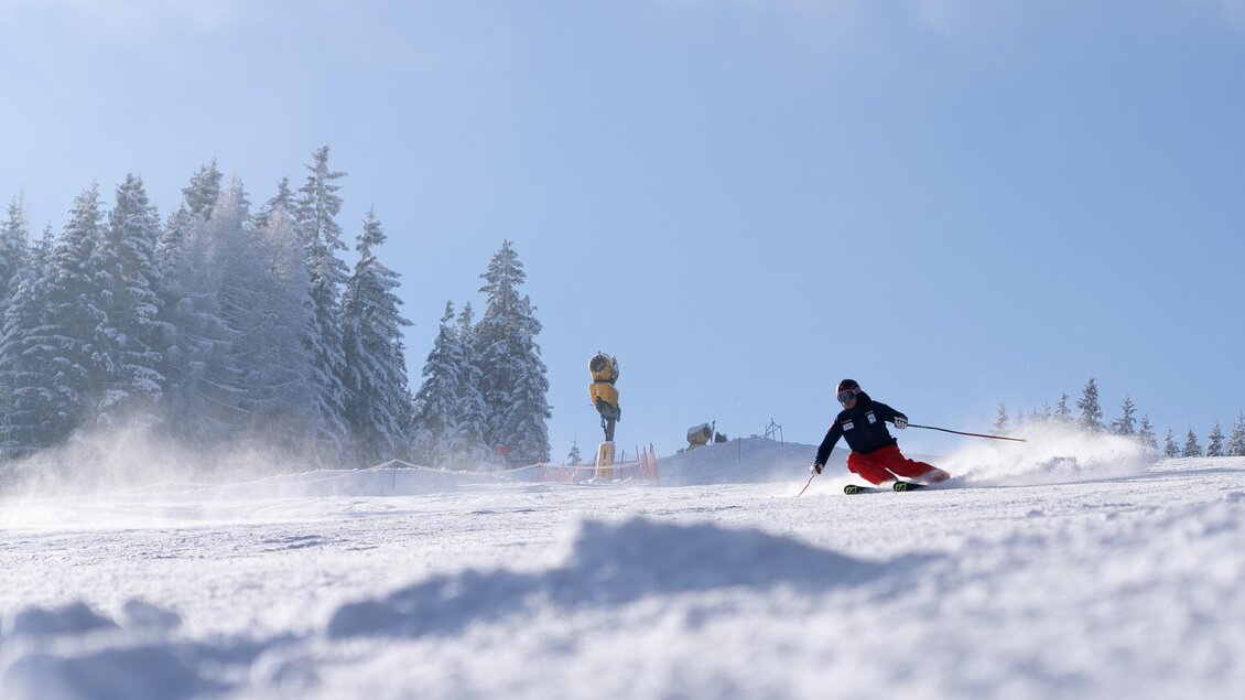 Ein Skifahrer fährt durch frischen, pulvrigen Schnee in der Berglandschaft. Im Hintergrund ist eine weitere Person zu sehen, umgeben von Bäumen und klarem blauen Himmel. | © Schilift Obdach