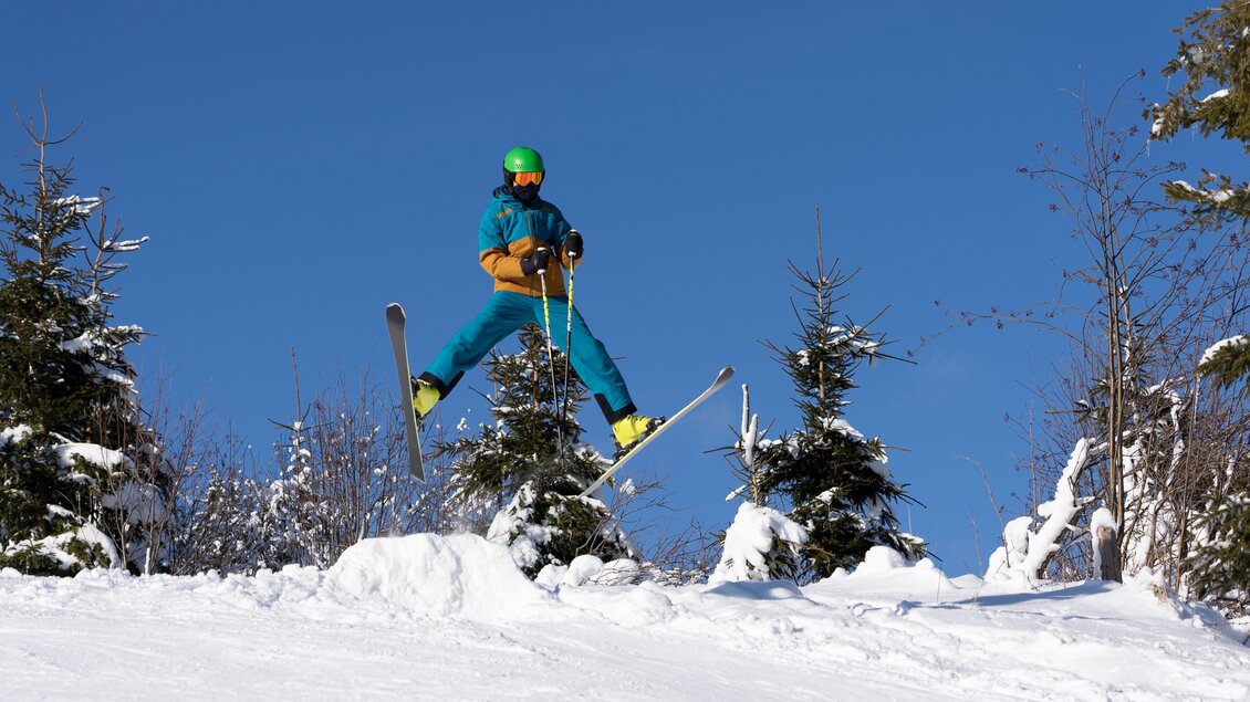 Ein Skifahrer springt über einen Schneeanhäufung in einer winterlichen Landschaft. Der Himmel ist klar und blau, umgeben von schneebedeckten Bäumen. | © Schilift Obdach
