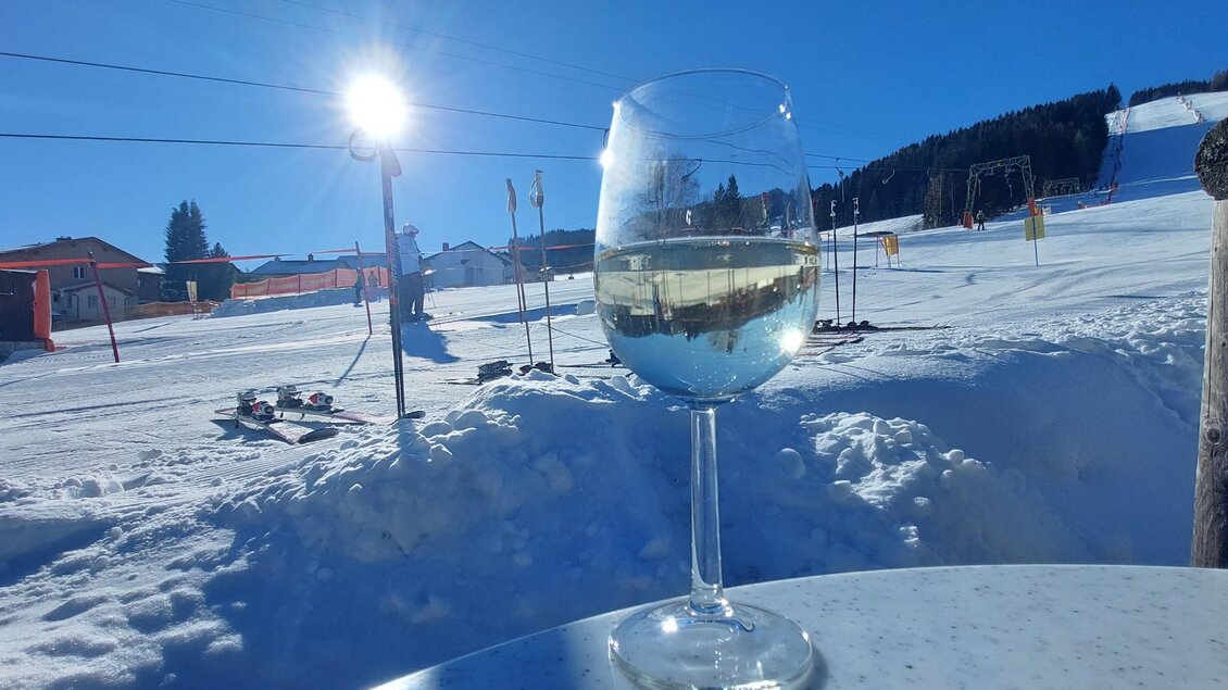 Ein Glas Weißwein steht auf einem Tisch im Schnee. Im Hintergrund sind die Skilifte und die klare blaue Himmel zu sehen. | © Schilift Obdach